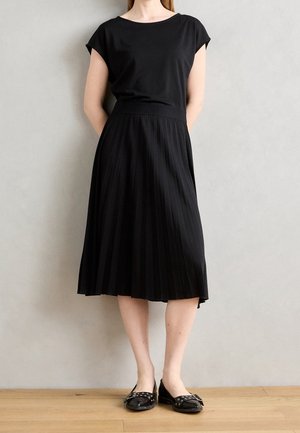 Sort kortærmet top kombineret med en sorte plisserede midi-nederdel, der fremhæver en glat tekstur. Modellen bærer sorte flade sko på et trægulv.