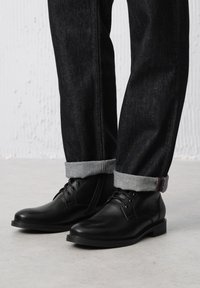 Czarne skórzane buty na sznurówki, gładka tekstura, zaokrąglony nos, w połączeniu z ciemnoniebieskimi dżinsami podwiniętymi przy mankietach, z jasnoszarym materiałem.