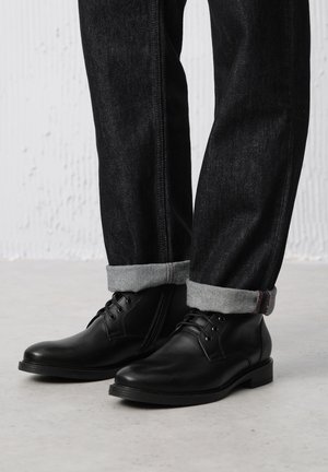 Czarne skórzane buty na sznurówki, gładka tekstura, zaokrąglony nos, w połączeniu z ciemnoniebieskimi dżinsami podwiniętymi przy mankietach, z jasnoszarym materiałem.