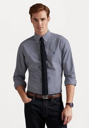 Blauw-wit ruitjesoverhemd met button-down kraag, lange mouwen opgerold, gecombineerd met een donkere gebreide stropdas en een bruine riem. Donkere jeans en een horloge zichtbaar.