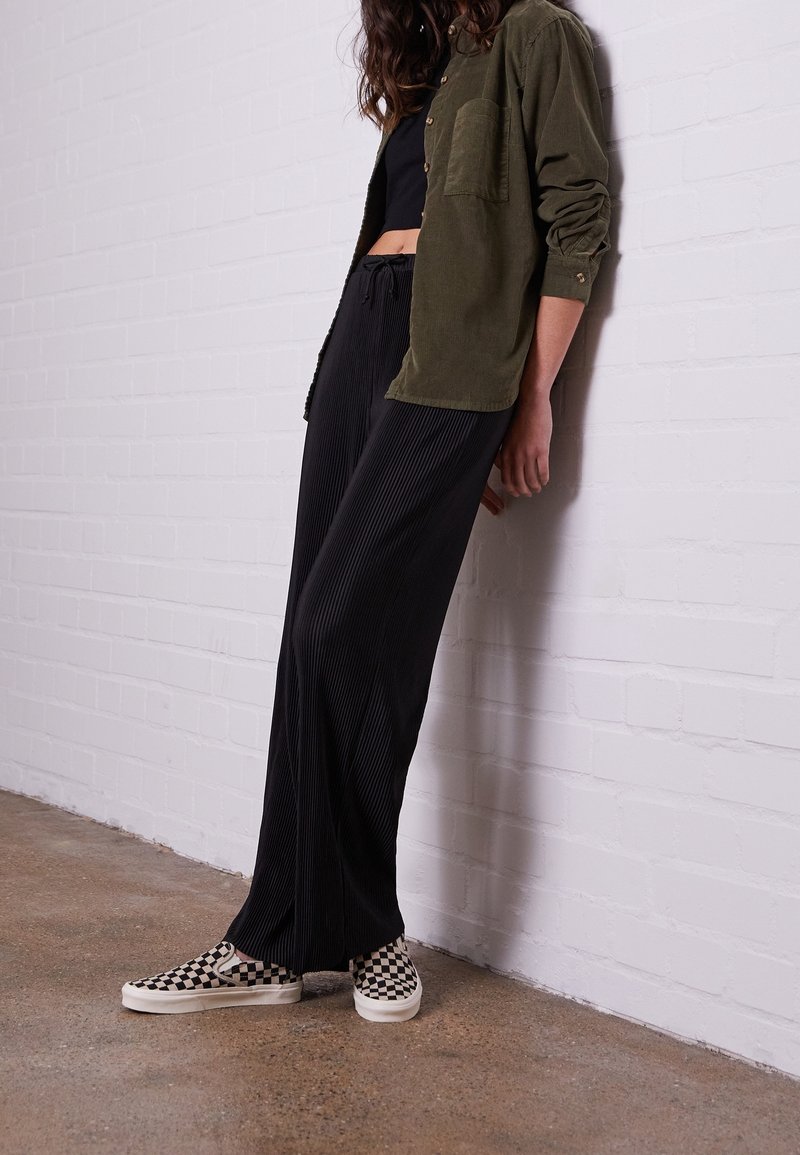 Personne appuyée contre un mur en briques blanches portant un pantalon large plissé sombre, une chemise boutonnée vert olive, un crop top noir et des chaussures à enfiler à carreaux.