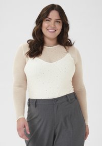 Langärmlige, durchsichtige beige Bluse mit silbernen Verzierungen über einer taillierten cremefarbenen Unterlage, kombiniert mit hochgeschnittenen grauen Hosen.