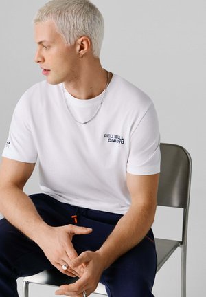 Weißes T-Shirt mit kurzen Ärmeln und dem in Marineblau aufgedruckten "RED BULL RACING"-Logo, kombiniert mit marineblauen Hosen mit orangefarbener Kordel. Dazu wird eine silberne Halskette getragen.