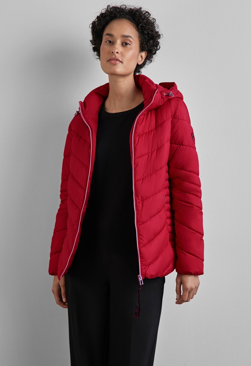 Rote Steppjacke mit einem gesteppten Design im Chevron-Muster, einem Reißverschluss, einer verstellbaren Kapuze und elastischen Bündchen. Schwarzes Oberteil darunter.