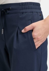 Marineblauwe broek van zachte stof met een elastische tailleband, trekkoordsluiting en zijzak. Kenmerken zijn het voorste plooidetail.