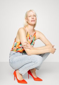 Top de alças com estampa floral colorida, combinado com jeans de cintura alta azul claro e sapatos de salto alto vermelhos brilhantes. O fundo é simples e neutro.