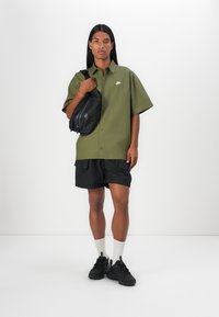 Green short-sleeve button-up shirt with Nike logo, paired with black shorts, black sneakers, and carrying a black sling bag.