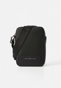 Melna taisnstūrveida plecu soma no fakturēta materiāla ar logotipa akcentu priekšpusē un regulējamu siksnu.