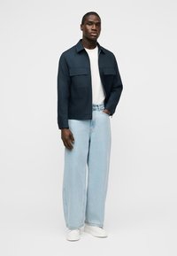 Άνδρας που στέκεται φορώντας ναυτικό τζάκετ πάνω από άσπρο πουκάμισο, ανοιχτό μπλε τζιν με φαρδιά μπατζάκια και λευκά αθλητικά παπούτσια, μπροστά από μια απλή φόντο.