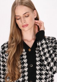 Svetr s černobílým houndstooth vzorem, pletenou texturou, V-výstřihem a kovovými knoflíky; dlouhé, vlnící se vlasy viditelné v rámu.