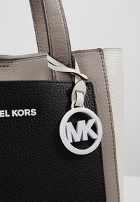 Sac cabas en cuir noir et gris avec une finition texturée, orné d'un charm circulaire blanc avec le logo « MK » et d'une plaque argentée avec le nom de la marque.