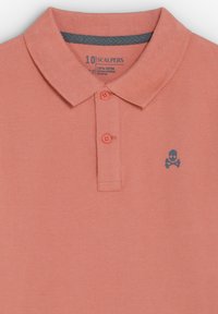 Polo color salmón con dos botones, cuello interior gris y pequeño logotipo oscuro de calavera y huesos cruzados en el lado izquierdo del pecho.