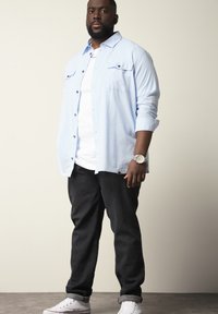 Lichtblauw button-up shirt over een wit t-shirt, gecombineerd met donkere jeans en witte sneakers. Zichtbare polshorloge om de linkerpols.