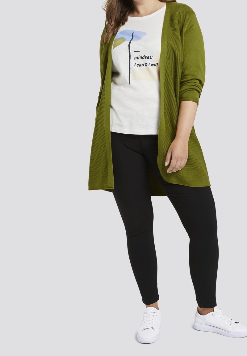 Cardigan largo color verde olivo sobre una camiseta blanca con un estampado gráfico, combinado con leggings negros y zapatillas blancas. Diseño de punto texturizado.