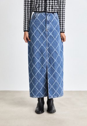 Femme portant une longue jupe en denim bleu avec un motif losange blanc et le texte "HUGO", associée à des bottes noires et une veste pied-de-poule noire et blanche.