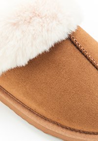 Pantofole in suede marrone con un morbido e leggero risvolto in finta pelliccia. Presentano cuciture tonali e una suola testurizzata. Aspetto liscio e caldo, adatte per l'uso indoor.