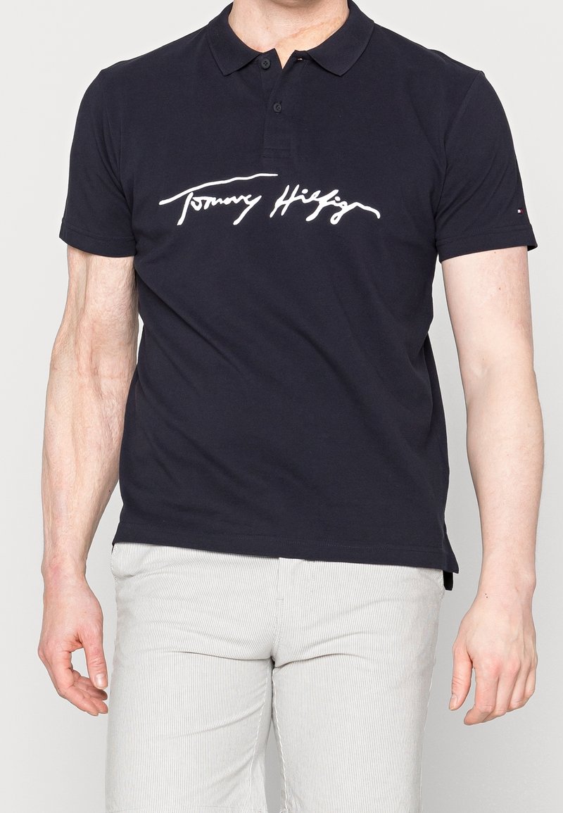 Homme portant un polo noir avec une écriture blanche "Tommy Hilfiger" sur la poitrine et un pantalon gris clair sur un fond uni.