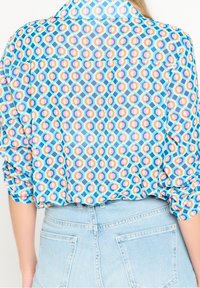 Camicia trasparente con bottoni caratterizzata da un motivo geometrico in blu, rosa e arancione. Maniche arrotolate e orlo curvo abbinati a pantaloni corti in denim.