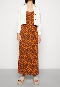 Robe longue marron à rayures tigre orange, associée à une veste courte en denim blanche ; complétée par des sandales compensées beiges.