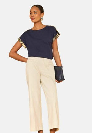 Nainen poseeraa laivastonsinisessä yläosassa, jossa on leopardikuvioiset hihat, kermaiset korkeavyötäröiset housut ja musta clutch-laukku kädessä.