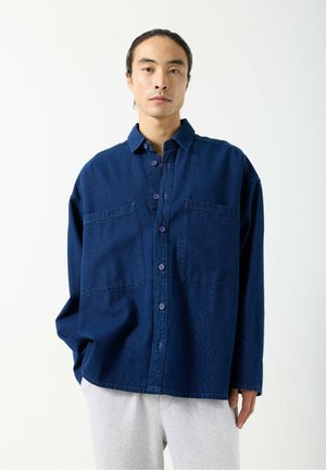 Jeune homme aux longs cheveux bruns attachés en queue de cheval basse, portant une chemise bleu foncé ample à boutons et un pantalon gris clair, debout devant un fond blanc.