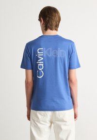 Blue cotton t-shirt featuring vertical white "Calvin Klein" text and logo on the back. Short sleeves and a crew neck design.