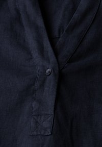 Camicia di lino blu navy con chiusura a bottone, design a scollo a V e dettagli di cucitura visibili, caratterizzata da una superficie morbida e testurizzata.