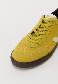 Geltoni zomšiniai sportbačiai su rudais guminiu padu, tekstūruotu detaliu ir kontrastingu baltu logotipu šone. Šnerves atitinkančios geltonos spalvos.