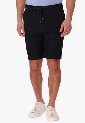 Uomo che indossa pantaloncini neri fino al ginocchio con coulisse e scarpe da ginnastica bianche, in piedi davanti a uno sfondo chiaro e uniforme.