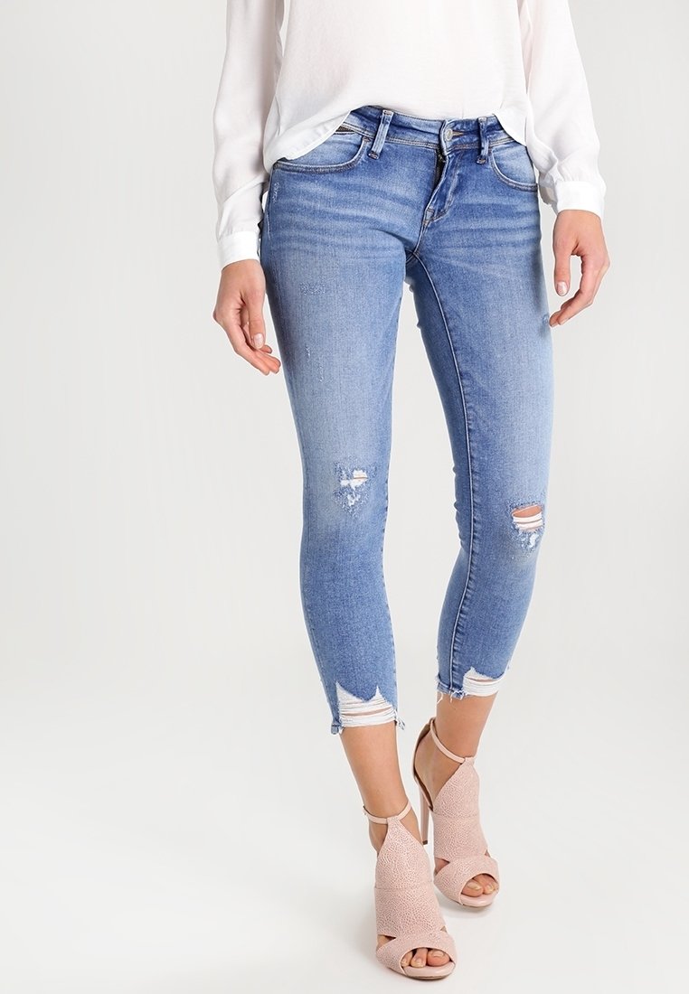 Jeans ajustados en denim de color azul claro con detalles desgastados, dobladillos deshilachados y un diseño de corte estrecho, combinados con tacones abiertos de color rosa pálido.
