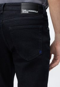Σκούρο τζιν από denim με μαύρο δερμάτινο έμβλημα με την ένδειξη «Karl Lagerfeld Jeans» στην πίσω μέση, φορεμένα από ένα άτομο με ορατό μπράτσο.