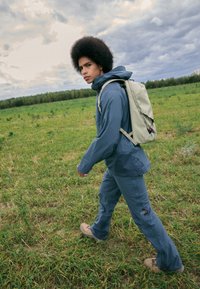 Hellblaues Outdoor-Jacket mit Kapuze, graue Hose und beiger Rucksack, der auf grünem Gras unter einem bewölkten Himmel geht.