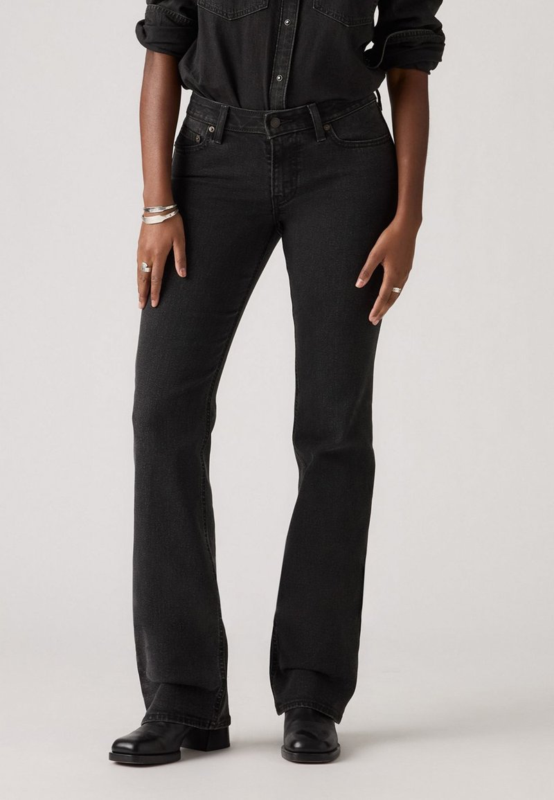 Jeans en denim noir évasés avec une taille haute, dotés de poches avant et d'une fermeture à bouton. Le tissu a une texture lisse et des détails de couture minimalistes.