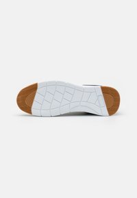 Biała gumowa podeszwa buta z teksturowanymi wzorami, z brązowym akcentem z gumy w okolicy pięty oraz segmentowanym wzorem zapewniającym przyczepność.