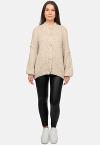 Cardigan beige a maglia con grandi bottoni, colletto a coste e maniche larghe, abbinato a leggings neri e sneakers bianche.