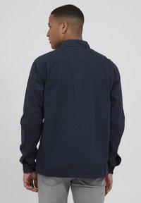 Camicia blu navy con bottoni, maniche lunghe, colletto piatto e orlo rotondo. Presenta due aperture laterali con bottoni sui polsini e una texture liscia.