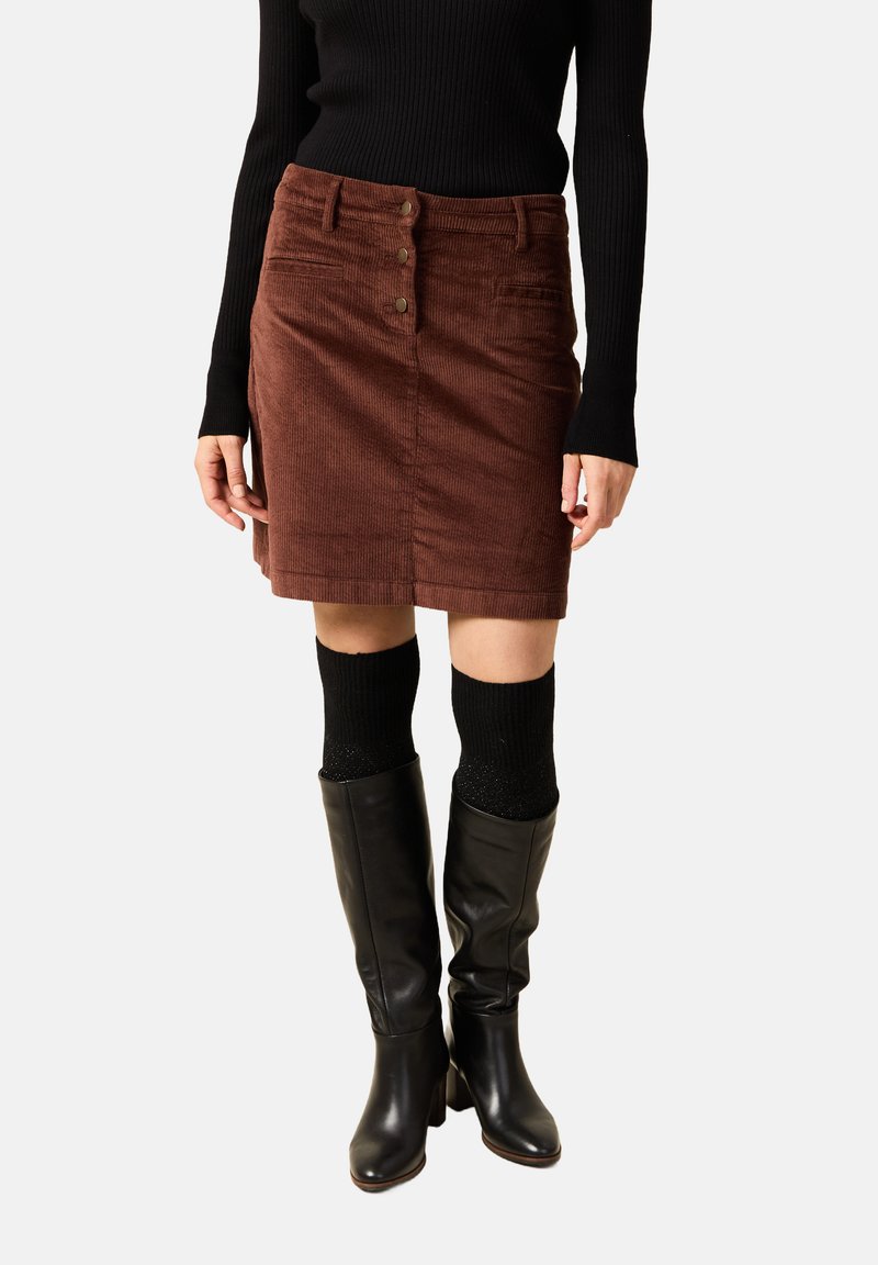 Ruskea korduroykangas hame, jossa on suora leikkaus, etunappikinoitus ja taskut, yhdistettynä polven korkeisiin mustiin nahkabootseihin ja mustiin teksturoituihin sukkiin.