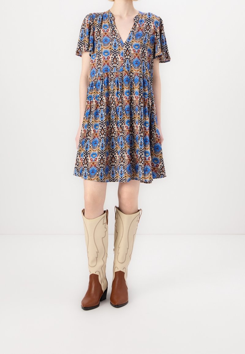 Femme portant une robe courte à motifs bleus, marron et noirs, assortie à de hautes bottes de cowboy en cuir crème et marron.