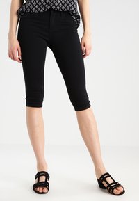 Čierne Capri nohavice so slim fit strihom, s hladkou textúrou a stredne vysokým pásom. Skombinované s čiernymi sandálmi na remienky.