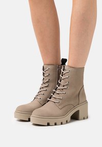 Beige ankle boots with a textured finish, lace-up design, silver hardware, and chunky, rubberized heels. Side zip for easy access.