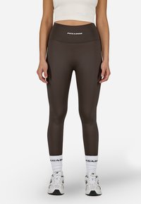 Barna, magas derekú leggings sima anyagból, a derékrészen logóval. Fehér zoknikkal és sportcipőkkel viselve.