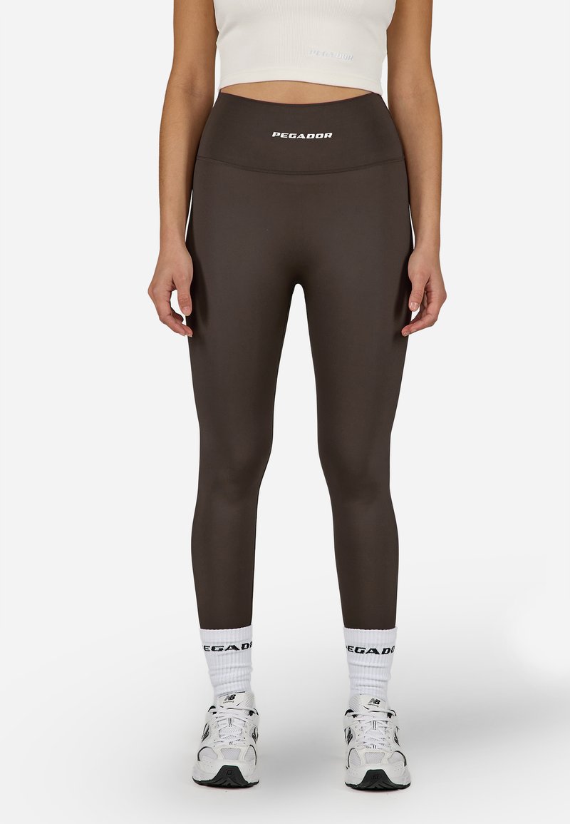 Barna, magas derekú leggings sima anyagból, a derékrészen logóval. Fehér zoknikkal és sportcipőkkel viselve.