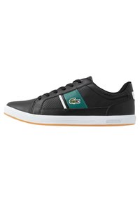 Zapatilla deportiva negra con parte superior sintética texturizada, panel de acento verde, franja horizontal blanca y suela de goma con una suela exterior marrón claro.