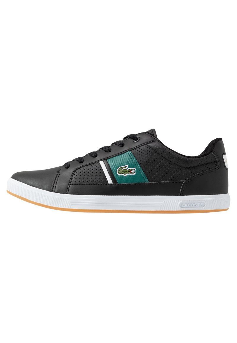Zapatilla deportiva negra con parte superior sintética texturizada, panel de acento verde, franja horizontal blanca y suela de goma con una suela exterior marrón claro.