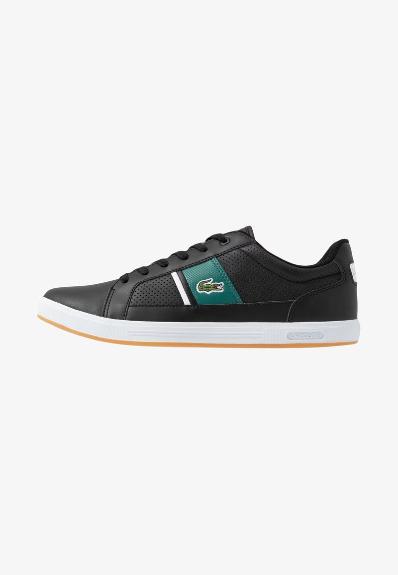Zapatilla deportiva negra con parte superior sintética texturizada, panel de acento verde, franja horizontal blanca y suela de goma con una suela exterior marrón claro.