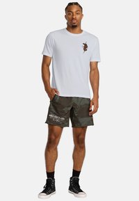 Jasnoszary t-shirt z małą zieloną grafiką na klatce piersiowej, w połączeniu z ciemnozielonymi szortami w kamuflażu z białym tekstem. Czarne trampki z wysoką cholewką.