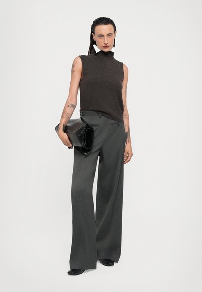 Brown sleeveless turtleneck top, gray wide-leg trousers, and a black clutch bag. Features tattoos and minimalistic accessories.
