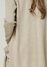 Beige, gestrickte Strickjacke mit langen Ärmeln, die eine Seitentasche mit einem Etikett enthält, auf dem steht "du bist perfekt." Weiche Textur und lässiges Design.