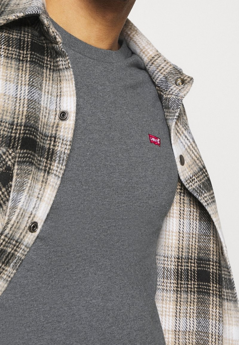 Personne portant un t-shirt gris Levi's sous une chemise beige et noire à carreaux ouverte sur un fond blanc.