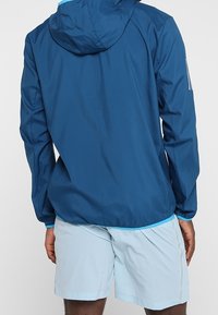 Chaqueta ligera azul marino con capucha y puños elásticos, combinada con pantalones cortos azul claro con un patrón texturizado.